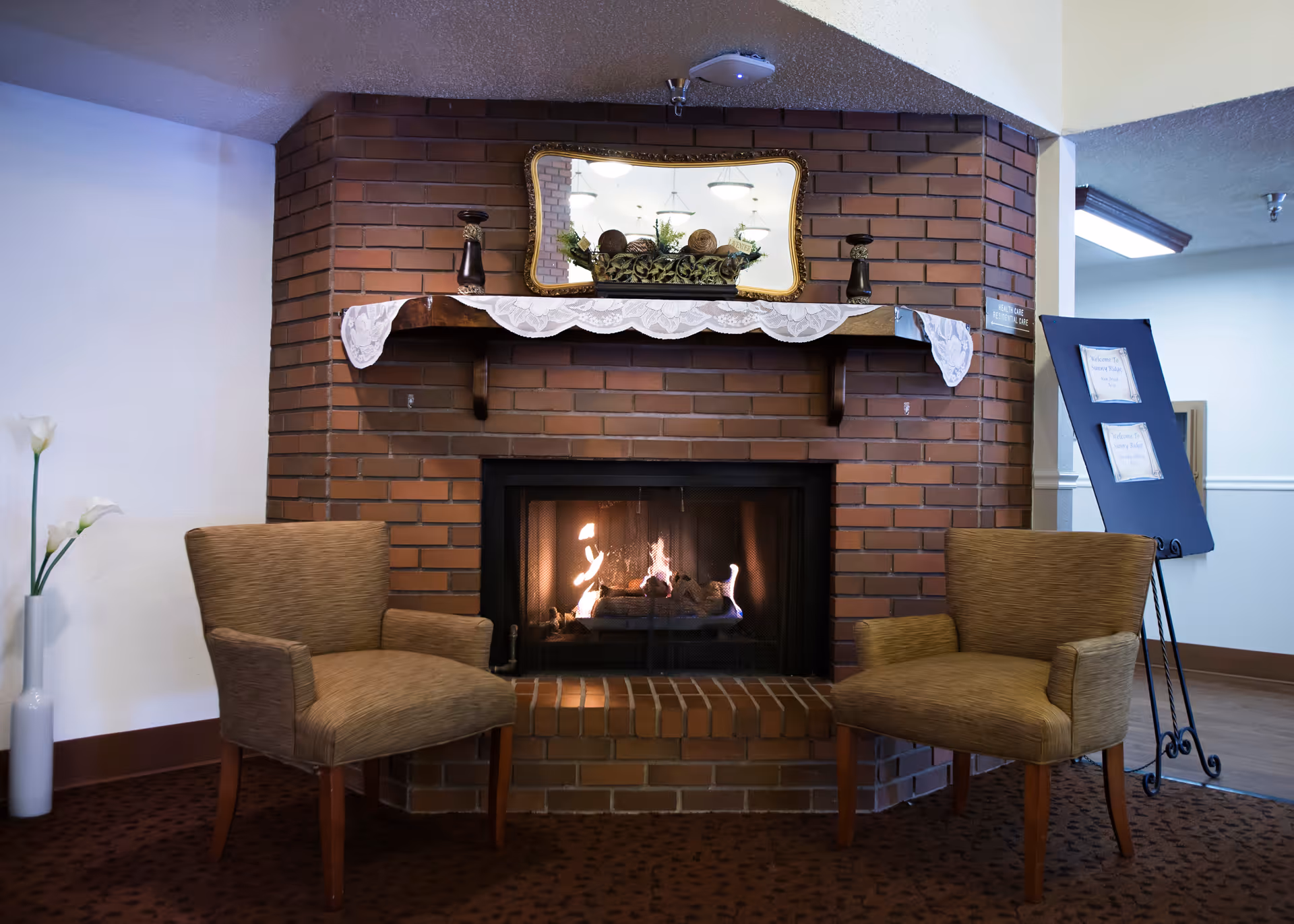 A cozy seating area with two brown upholstered chairs facing a brick fireplace with a fire burning. Above the fireplace is a wooden mantel decorated with a lace runner, two candle holders, and a decorative arrangement in front of a mirror. To the left is a tall white vase with white flowers, and to the right is a blue stand with framed notices.