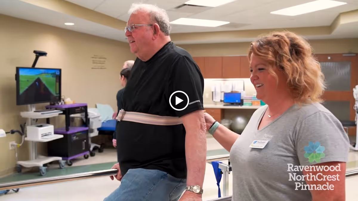 An elderly man wearing a black shirt and jeans is supported by a female caregiver as he walks between parallel bars in a rehabilitation or therapy room. The caregiver is smiling and holding the man's arm for support. In the background, there is medical equipment, a computer monitor, and a TV screen displaying a virtual track scene.