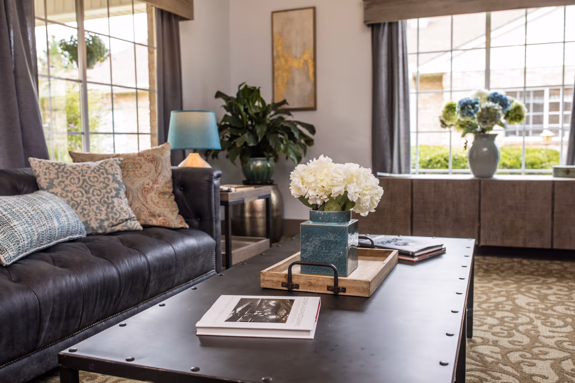 A cozy living room area with a black leather sofa adorned with patterned cushions, a black metal coffee table with a wooden tray holding a blue vase filled with white flowers, and a side table with a blue lamp. Large windows with gray curtains let in natural light, and a cabinet with a vase of blue and green flowers is visible in the background.