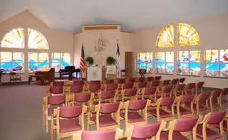 Interior of a chapel-like meeting room with rows of red-cushioned chairs facing a lectern and large arched windows.