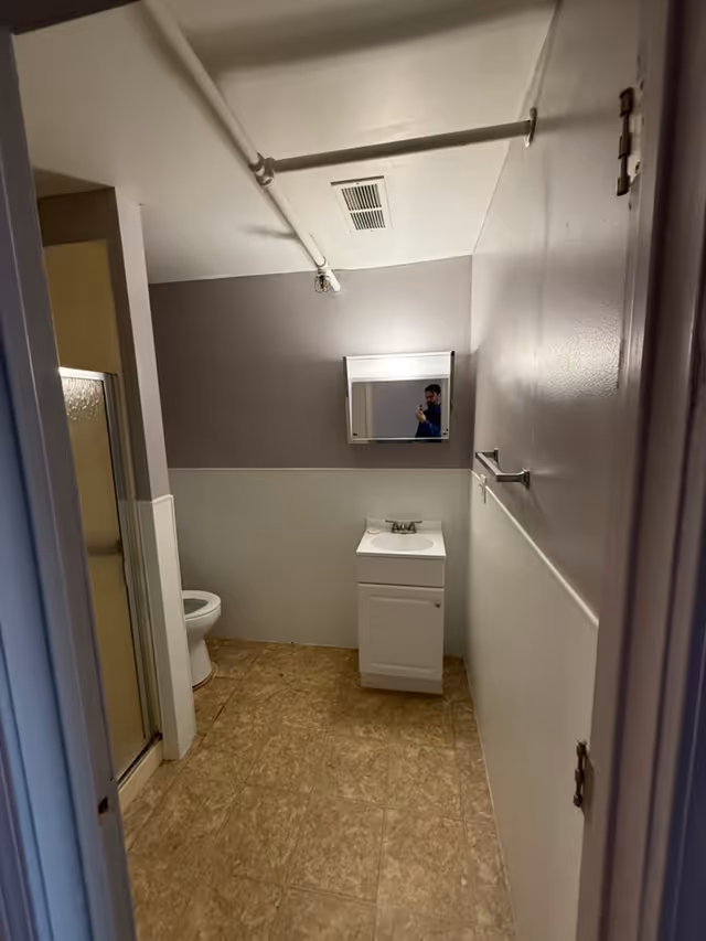 A small bathroom with beige tiled floor, a white toilet partially visible on the left, a white sink cabinet with a faucet in the center, and a mirrored medicine cabinet above the sink. The walls are painted gray on the upper half and white on the lower half. There is a towel rack on the right wall and exposed white pipes on the ceiling.