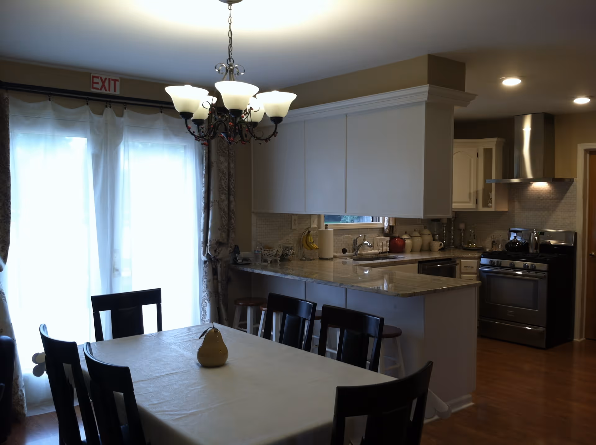 Interior view of a dining area and kitchen in a senior living facility. The dining area features a rectangular table covered with a white tablecloth and surrounded by six dark wooden chairs. A decorative pear-shaped object is placed on the table. Above the table hangs a chandelier with multiple light fixtures. The kitchen has white cabinets, a marble countertop, a stainless steel stove with a range hood, and various kitchen items on the counter. Sheer curtains cover the glass doors in the background, with an exit sign above them.