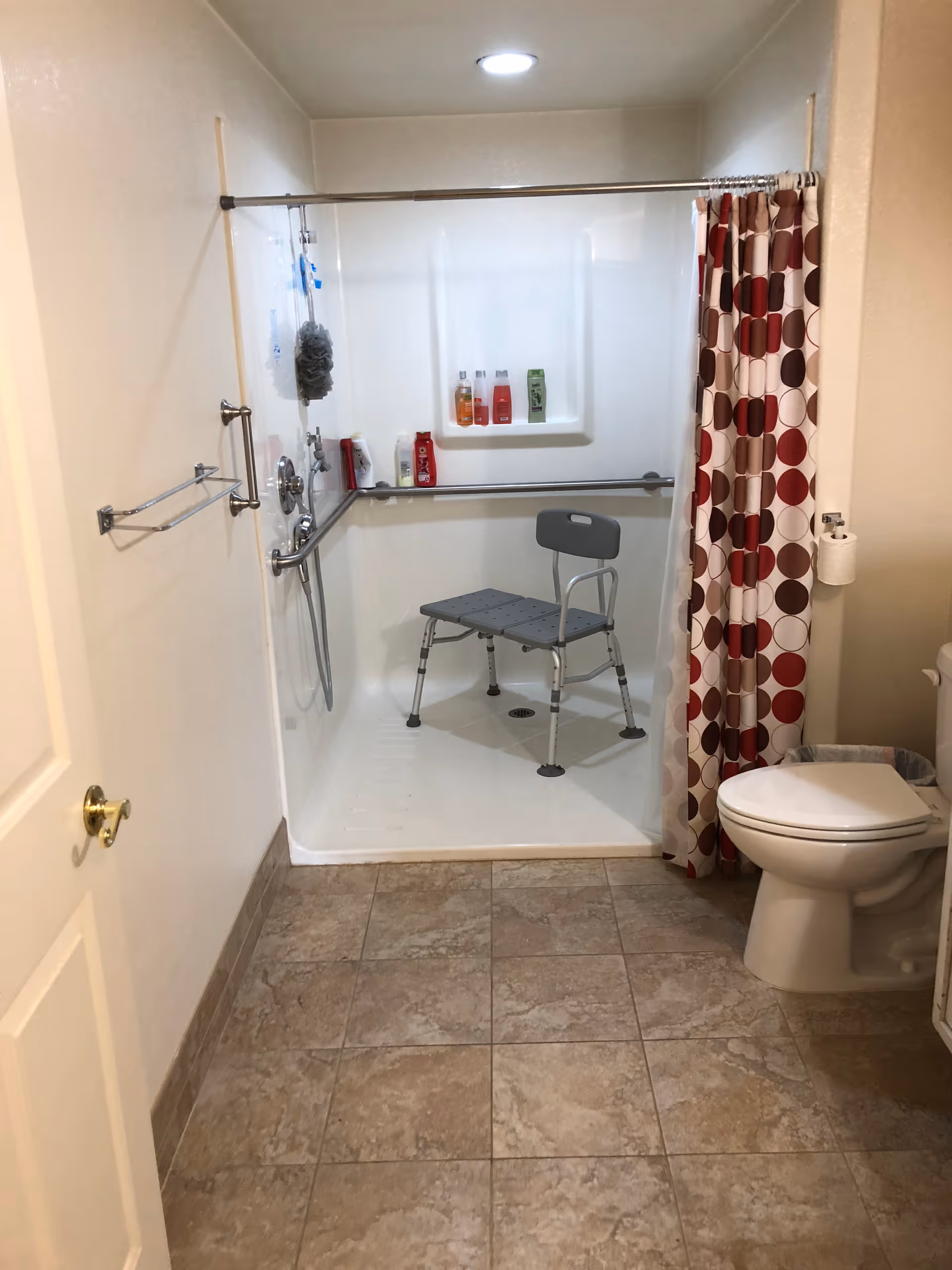 A bathroom with a tiled floor, a white toilet, and a shower area with a shower chair. The shower has grab bars, a handheld showerhead, and several bottles of toiletries on a built-in shelf. A shower curtain with a red, brown, and beige circular pattern is partially drawn.