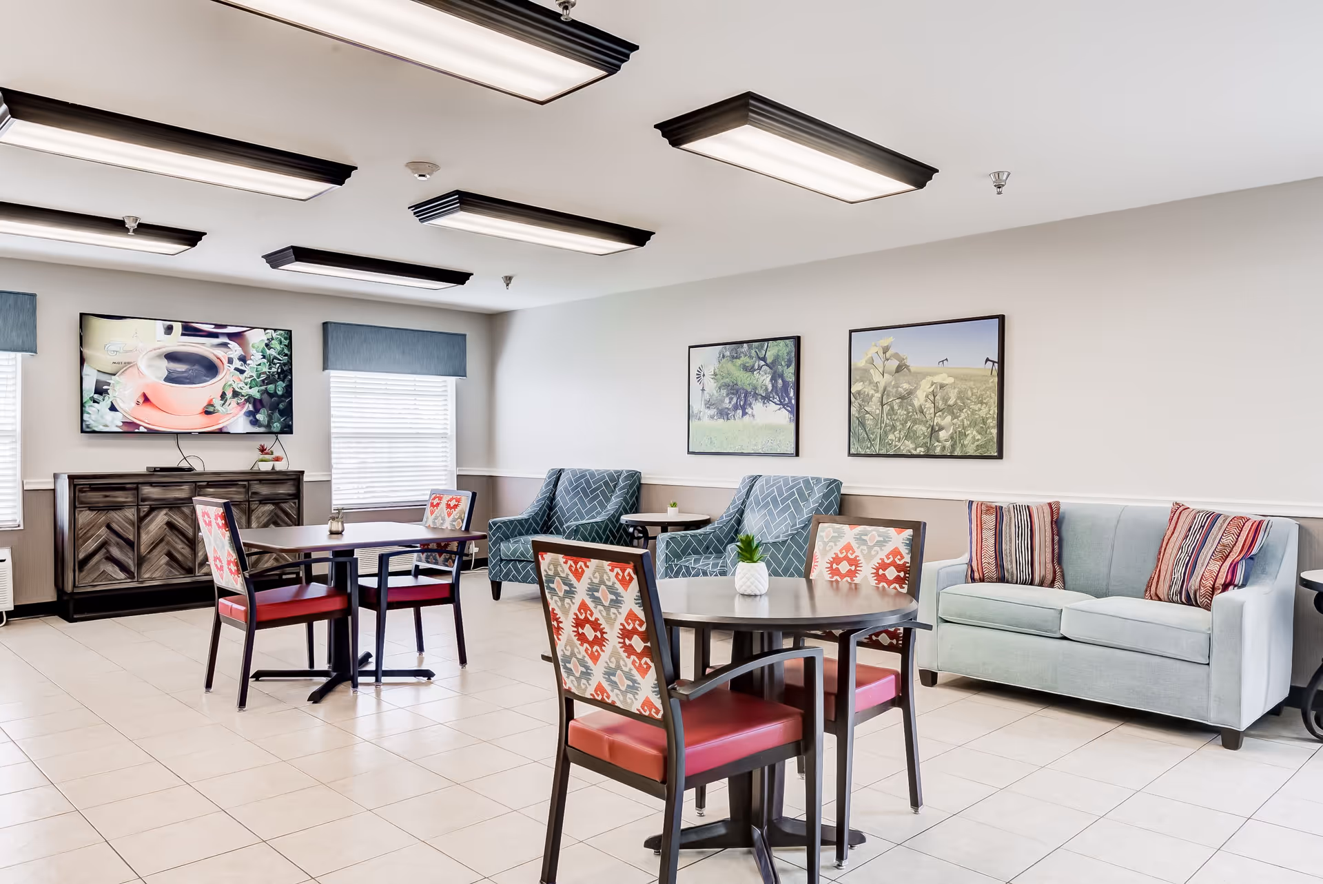 Bright communal dining and lounge area with tables and patterned chairs, a sofa and armchairs, wall art, and a TV.
