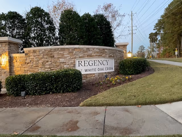 Stone entrance sign reading "Regency at White Oak Creek" set in a landscaped stone wall beside a sidewalk.
