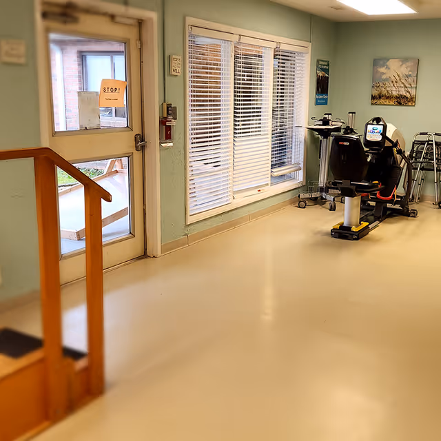 Interior view of a rehabilitation center room with a stationary exercise bike, walkers, a window with blinds, a door with a stop sign, and a small wooden handrail on the left side.