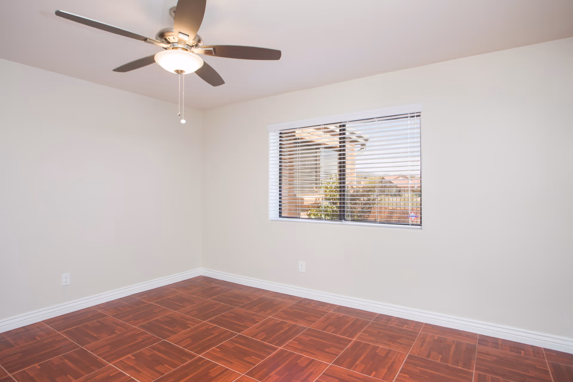 Empty room with white walls, a ceiling fan with light, a window with blinds showing an outdoor view, and reddish-brown tiled flooring.