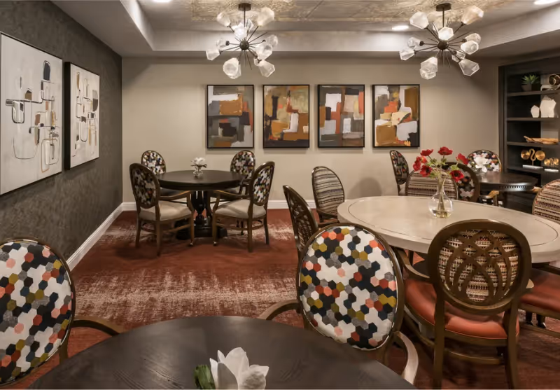 Interior view of a dining room with multiple round tables and chairs featuring colorful patterned upholstery. The room has modern light fixtures on the ceiling, abstract artwork on the walls, and a vase with red flowers on one of the tables.