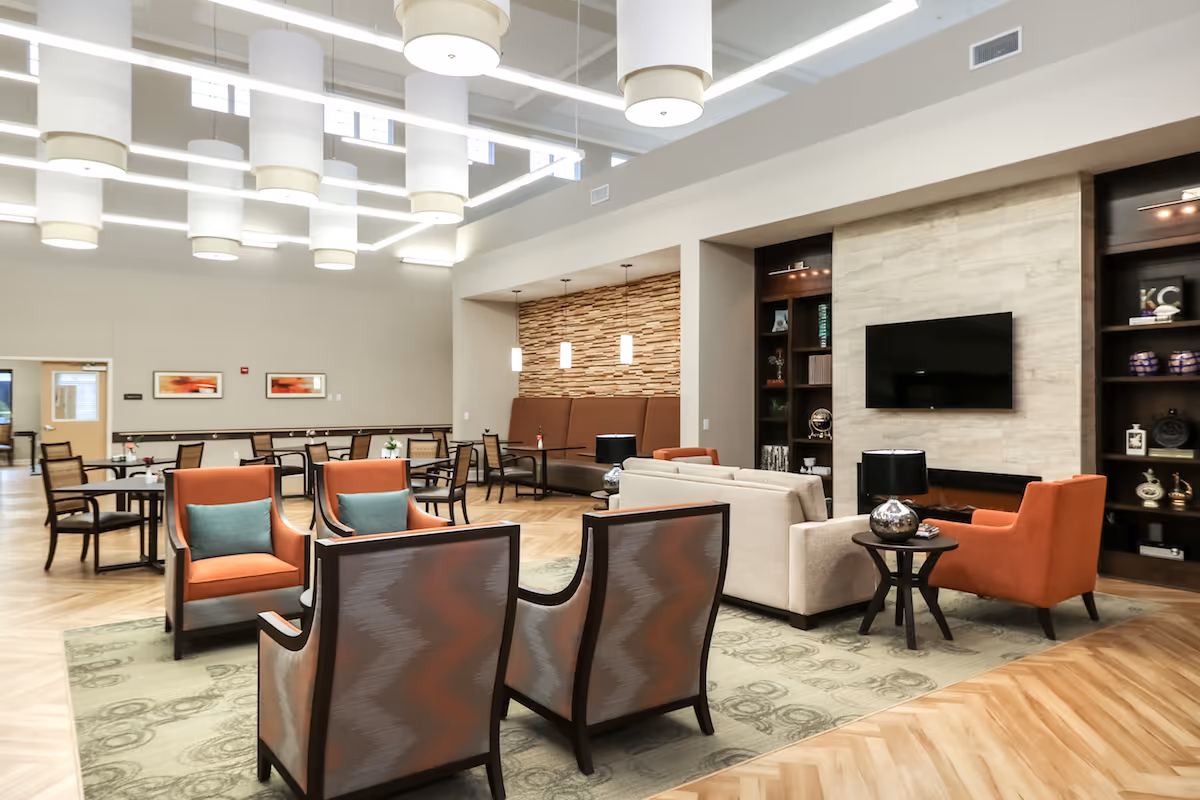 A spacious and well-lit common area in a senior living facility featuring a seating arrangement with orange and patterned armchairs, a beige sofa, a wall-mounted TV above a modern fireplace, and built-in shelves with decorative items. The room has wooden flooring, a green patterned rug, and multiple tables and chairs in the background. The ceiling has large cylindrical pendant lights and rectangular light fixtures.