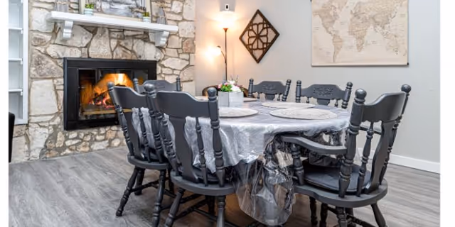 A dining area with a rectangular table covered with a plastic tablecloth and six black wooden chairs around it. There is a stone fireplace with a fire burning on the left side, a wall-mounted decorative piece, and a world map on the wall. The floor is wood-style laminate.