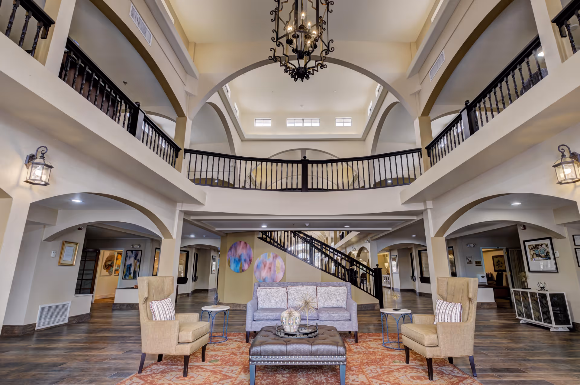 Spacious and elegant senior living facility common area with high vaulted ceilings, a large chandelier, and a second-floor balcony overlooking the space. The room features a seating arrangement with a sofa, two armchairs, a cushioned ottoman, and side tables on a patterned rug. The walls have decorative art pieces and the floor is wood with a warm finish.