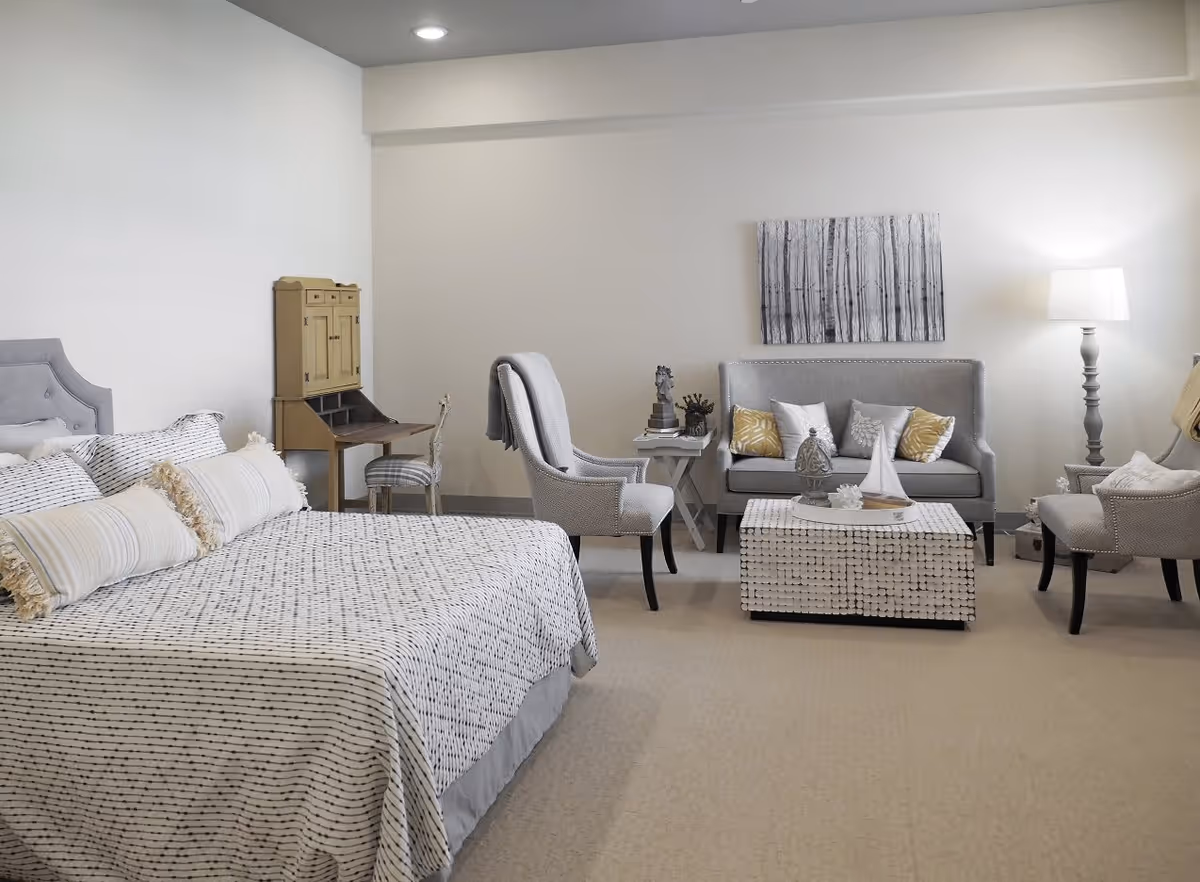 A spacious bedroom with a large bed covered in a patterned bedspread and multiple pillows. To the right, there is a sitting area with a gray loveseat adorned with decorative pillows, two upholstered armchairs, a white textured coffee table with decorative items, and a floor lamp. A small wooden desk with a chair is positioned against the far wall. The walls are light-colored, and a black and white forest-themed artwork hangs above the loveseat.