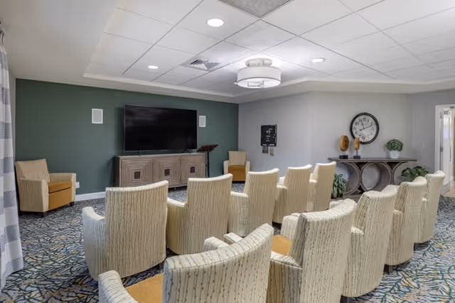 Small community media room with rows of upholstered chairs facing a wall-mounted TV and console, decorative clock and side tables.