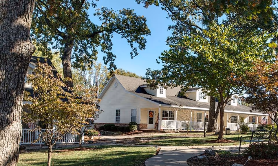 Front exterior of a single-story senior living building with white siding, porches, trees, and a curved walkway.