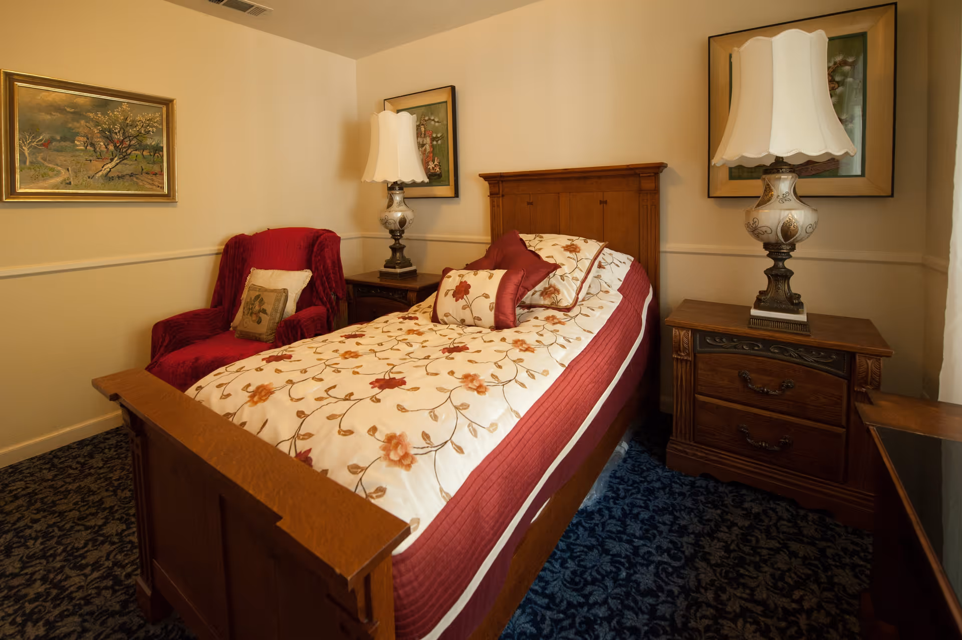 A cozy bedroom with a wooden bed featuring floral bedding in shades of cream and red. There is a red upholstered armchair with a decorative pillow to the left of the bed. Two wooden nightstands flank the bed, each with an ornate lamp and framed artwork hanging above. The room has beige walls and a dark patterned carpet.