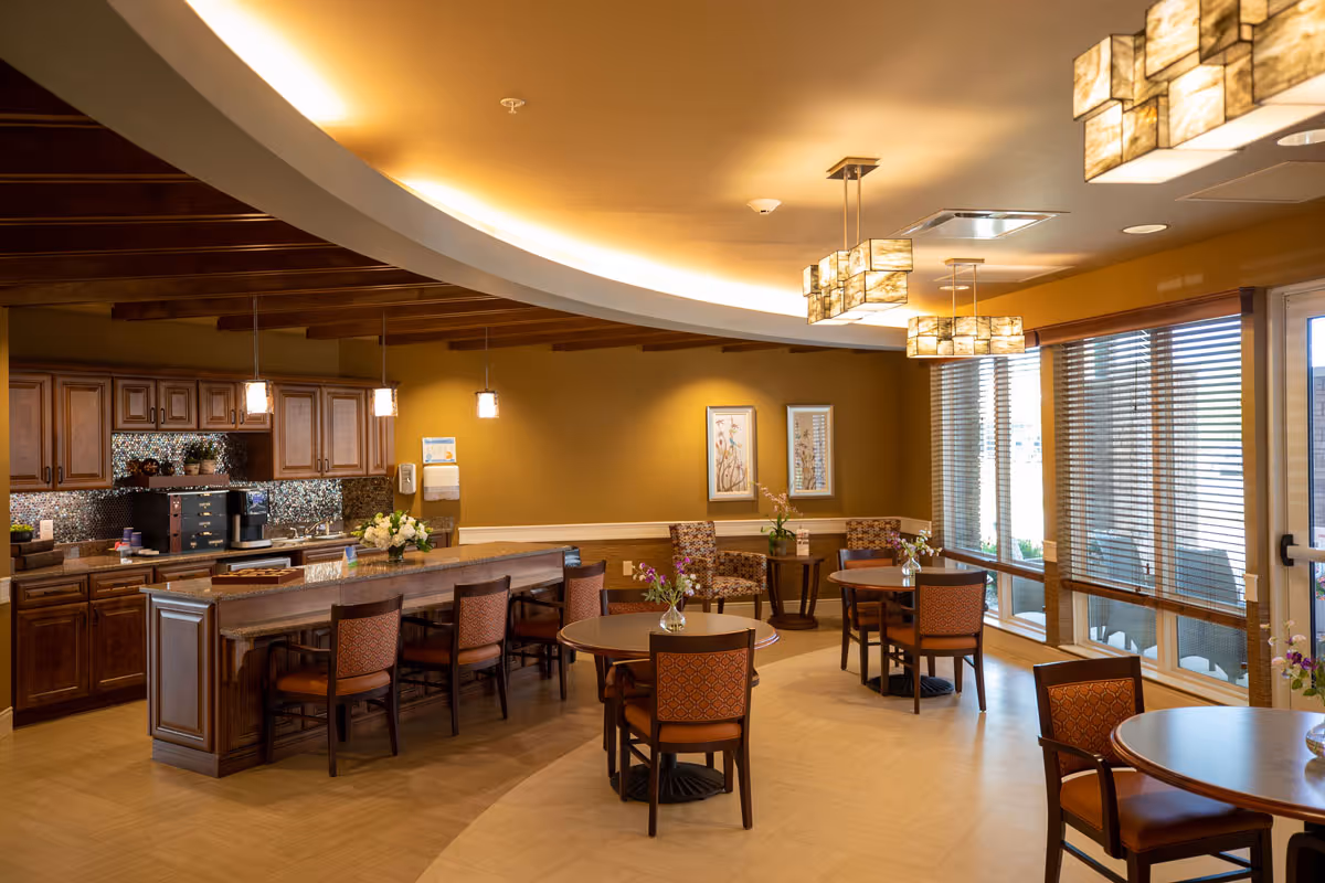 Warm, well-lit communal dining area with round tables, chairs, a long serving counter, and large windows.