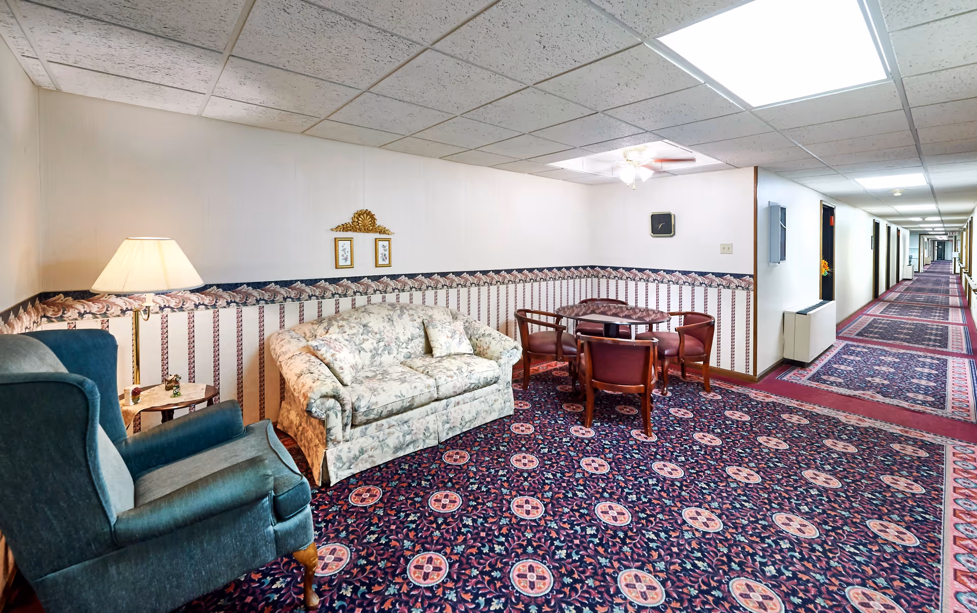 A sitting area in a senior living facility hallway featuring a floral patterned couch, a green upholstered armchair, a small round table with four wooden chairs, a floor lamp, and decorative wall art. The area has a patterned carpet and wallpaper with a floral border. The hallway extends to the right with multiple doors and exit signs visible.