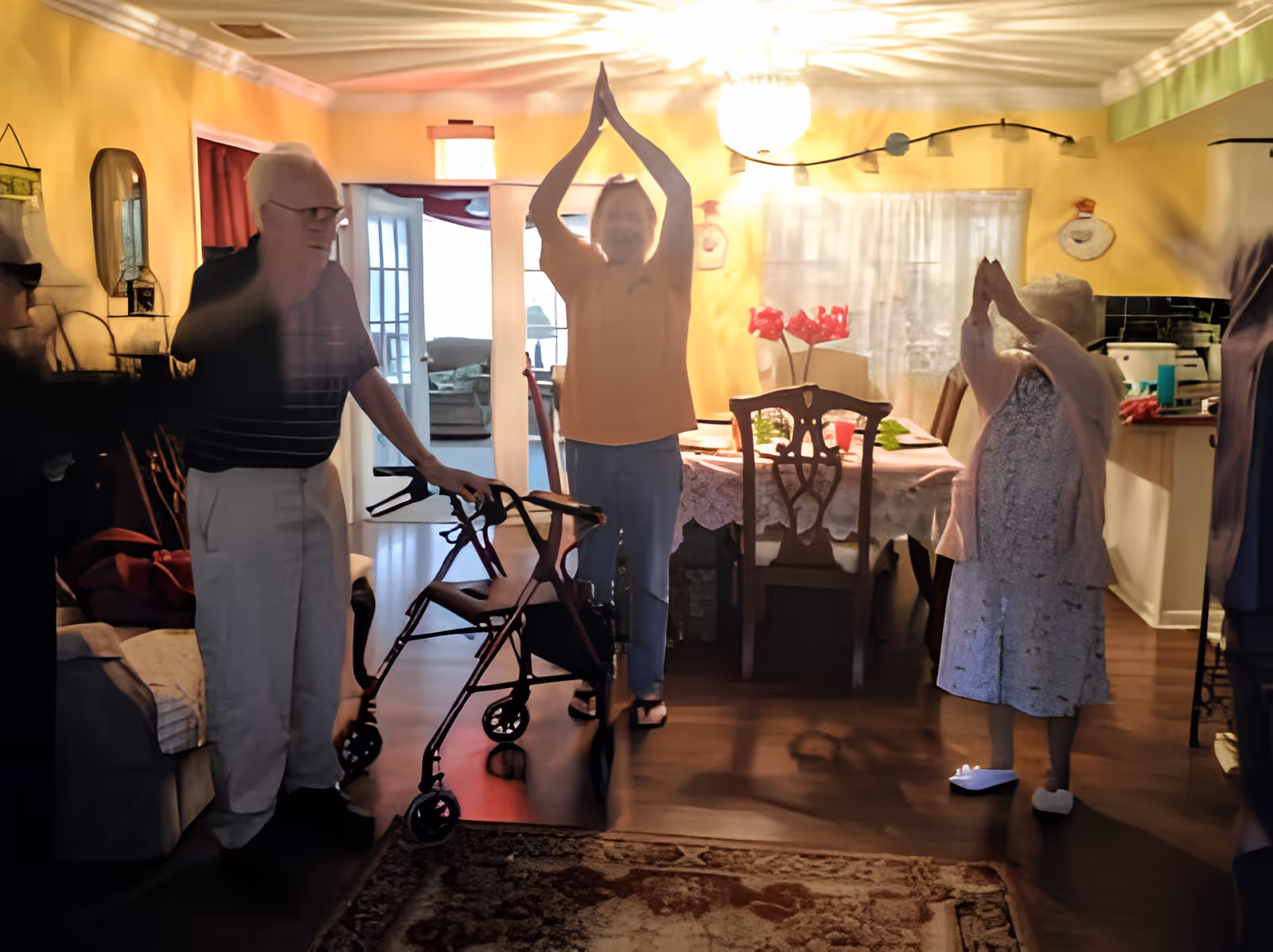 Three elderly individuals in a warmly lit living and dining area. One man with white hair and glasses is standing with a walker near a couch. Two women are standing with their arms raised above their heads, appearing to be engaged in a stretching or exercise activity. The room has yellow walls, a dining table with a lace tablecloth and chairs, a chandelier, and a kitchen area visible in the background.