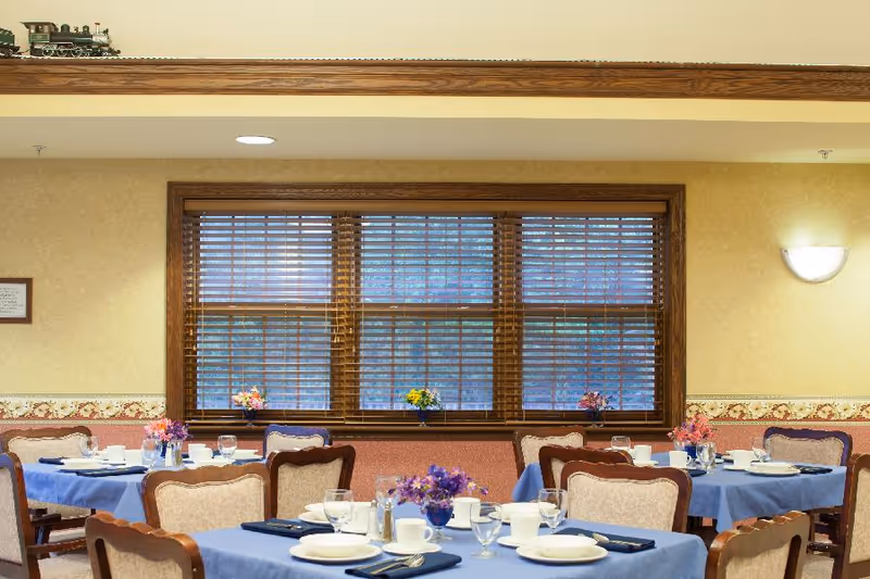 A dining room with several tables covered in blue tablecloths, each set with white plates, cups, glasses, silverware, and napkins. There are floral centerpieces on each table. The room has beige walls with a floral border and a large window with wooden blinds. A model train is displayed on a ledge near the ceiling.