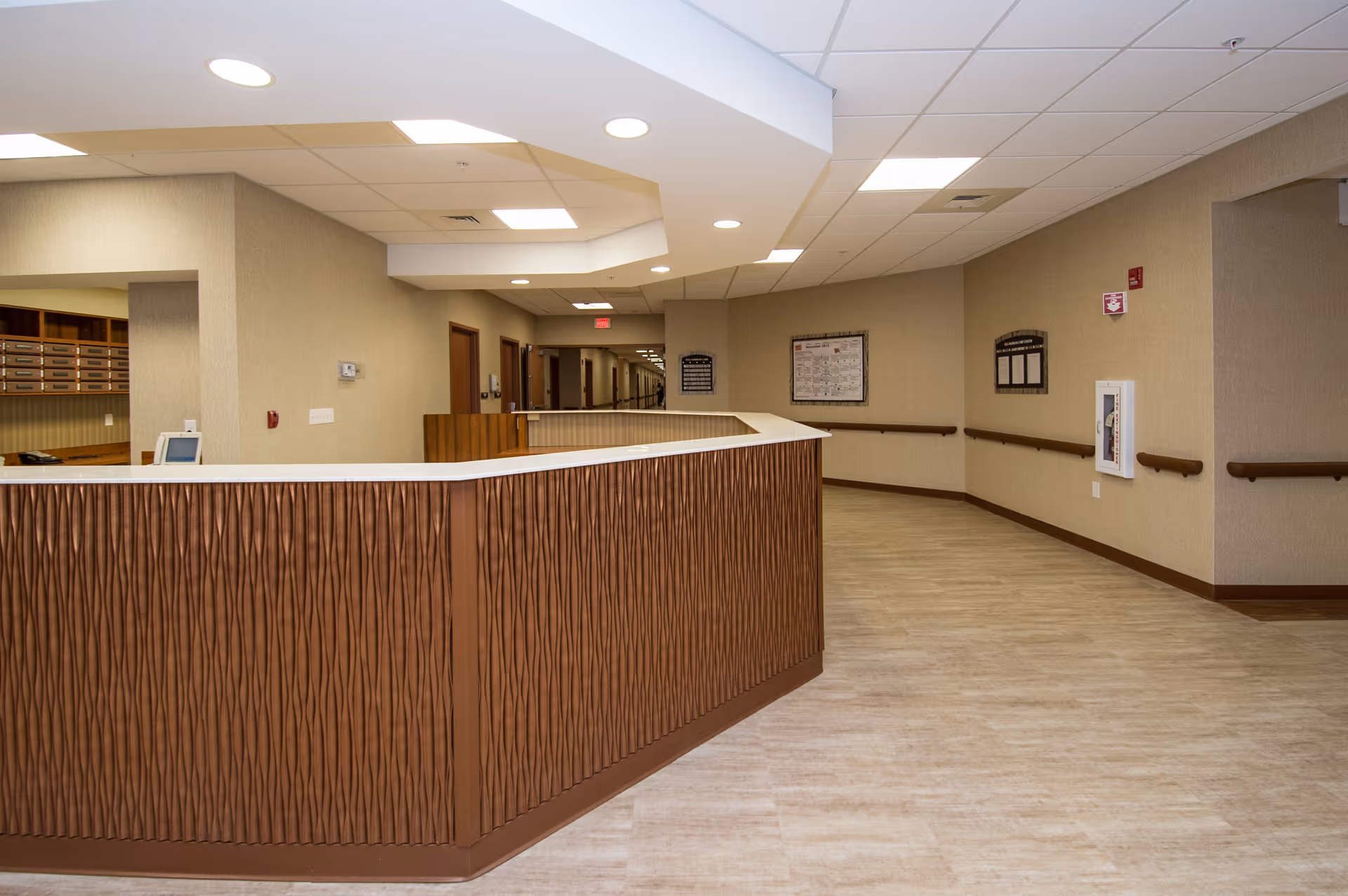 Curved wooden reception/nurse station and empty hallway inside a senior living facility with beige walls and overhead lighting.