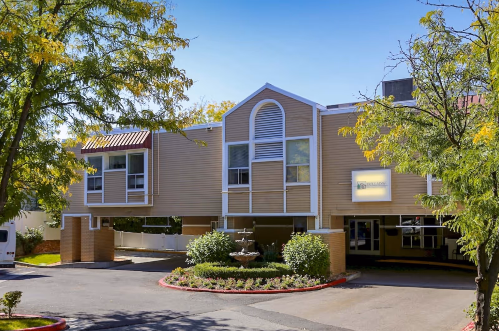Exterior view of Holladay Healthcare Center building with beige siding, white trim, and large windows. The building is elevated on brick pillars with a driveway underneath. There is a small landscaped area with a multi-tiered fountain and greenery in front. Trees with green leaves frame the scene under a clear blue sky.