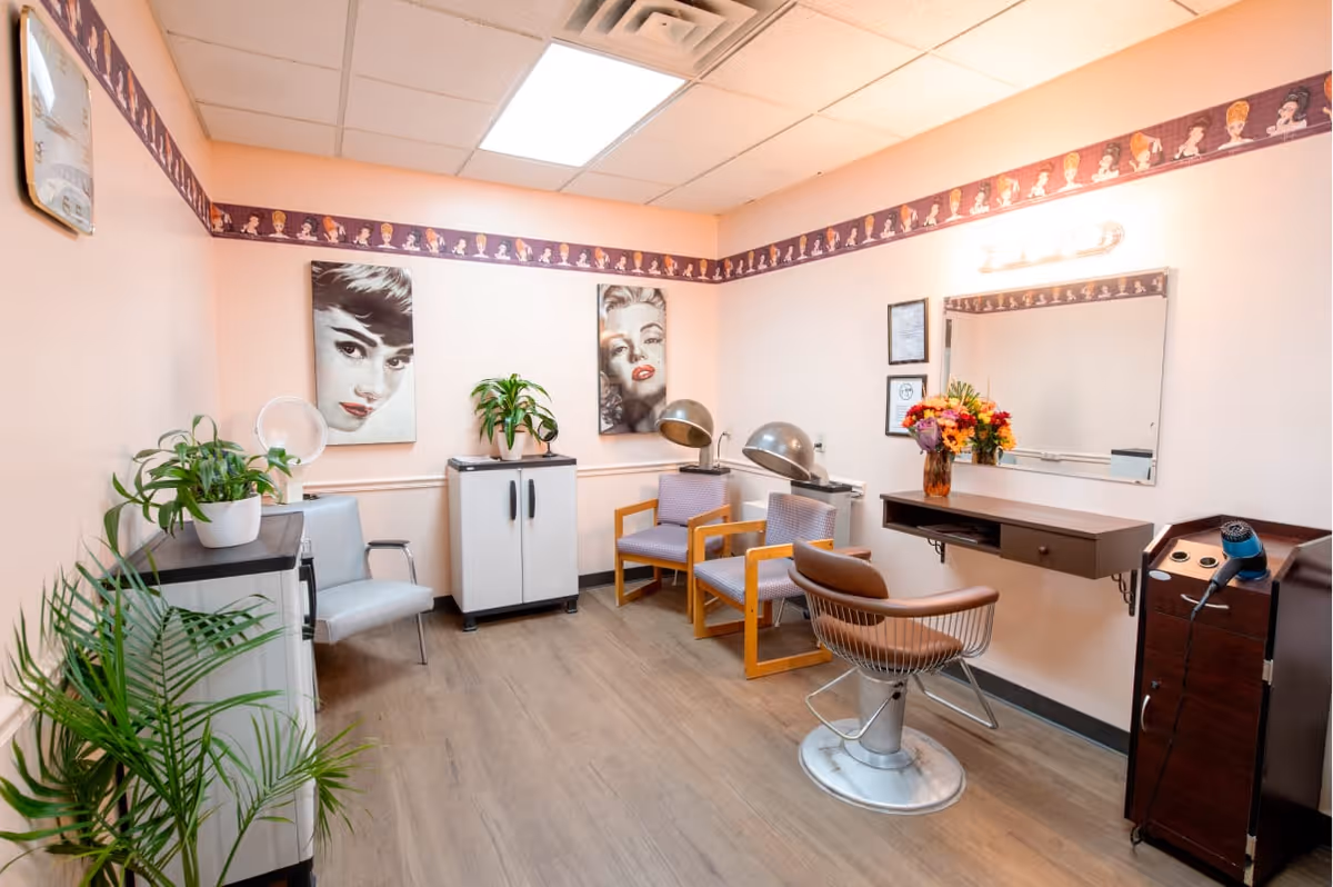A small salon room with light pink walls and wood flooring. The room contains a salon chair in front of a wall-mounted mirror with a floating shelf holding a vase of colorful flowers. There are two hair drying chairs with hooded hair dryers, two framed black and white portraits of women on the wall, several potted plants, and a hair dryer on a stand. The ceiling has fluorescent lighting and a decorative border featuring illustrated women's faces runs along the top of the walls.