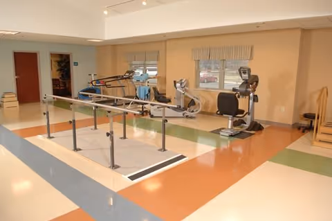 Bright rehabilitation room with parallel walking bars and various exercise machines near windows.