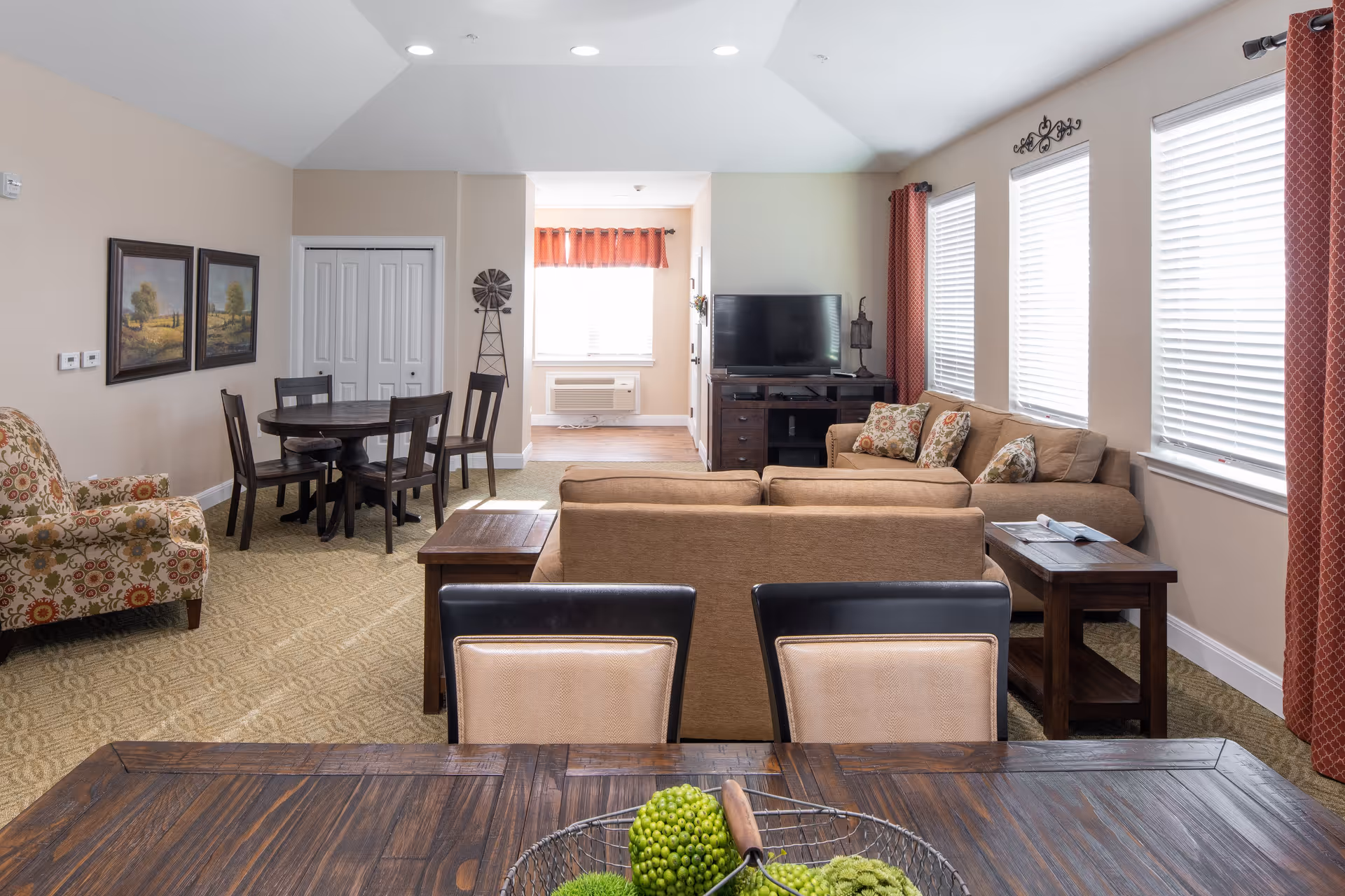 A bright and spacious living room area in an assisted living facility featuring a beige sofa with patterned cushions, a wooden TV stand with a flat-screen TV, a round dining table with four chairs, a patterned armchair, and large windows with white blinds and red curtains. The room has beige walls, carpeted flooring, and decorative wall art.