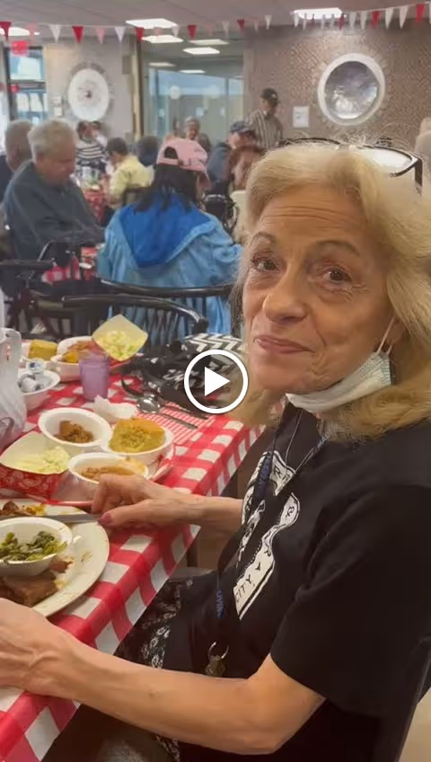 An elderly woman with blonde hair wearing a black shirt and a face mask under her chin is sitting at a table with a red and white checkered tablecloth. She is eating a meal with various dishes in front of her. In the background, several other elderly people are seated at tables in a dining area decorated with small triangular flags hanging from the ceiling.