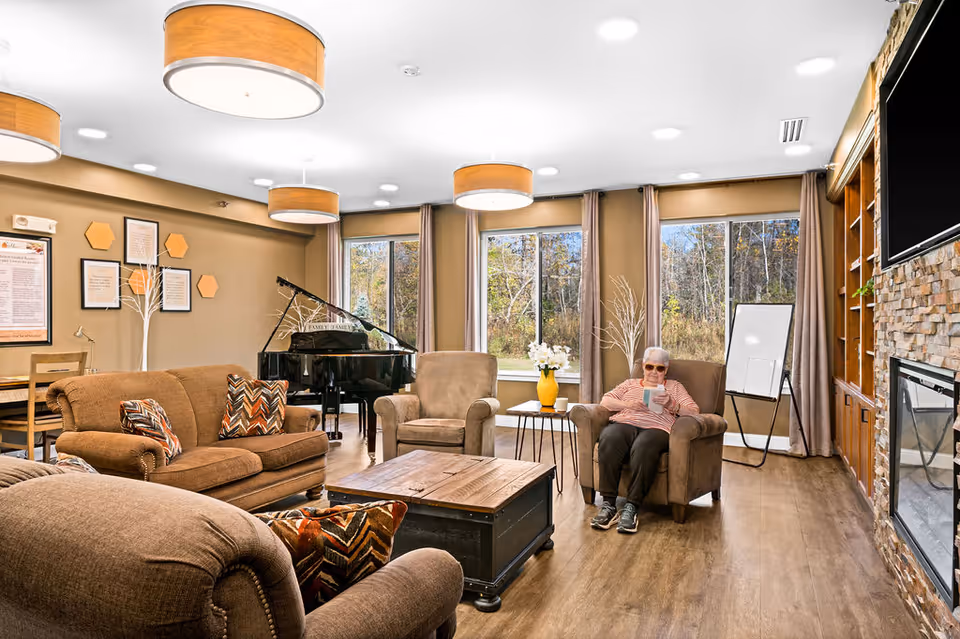 A cozy living room in an assisted living facility with brown upholstered sofas and armchairs, a wooden coffee table, large windows with beige curtains showing a wooded outdoor view, a black grand piano, a fireplace with a stone surround and a mounted TV above it, and a senior woman sitting in an armchair reading a book.