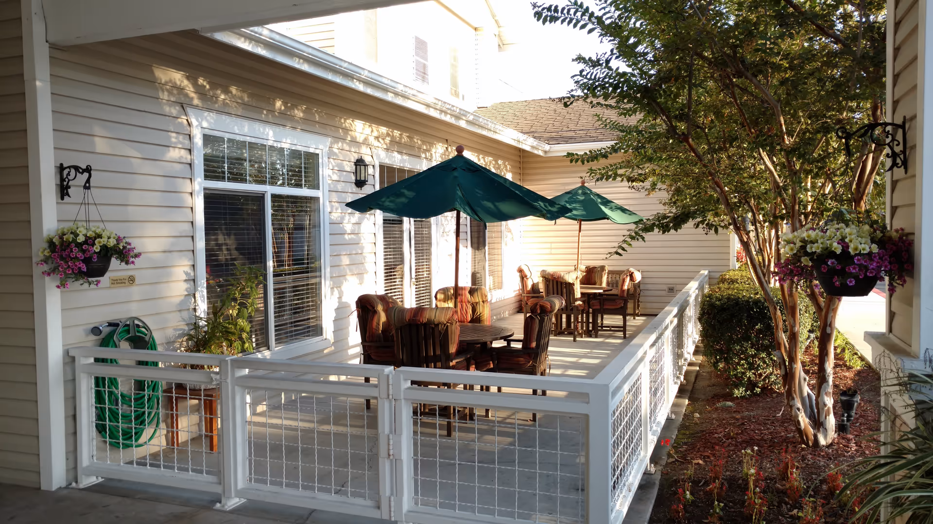 Outdoor patio area at Discovery Commons Whittier with several tables and chairs under green umbrellas. The patio is enclosed by a white fence and surrounded by plants and trees. The building exterior is light-colored with windows and hanging flower pots.