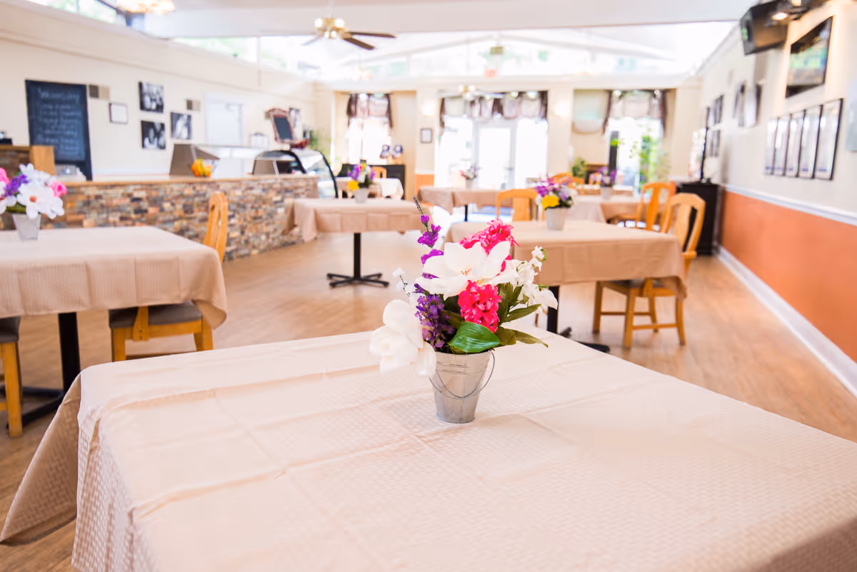Interior view of a dining room with several tables covered in beige tablecloths, each decorated with a small metal bucket holding colorful artificial flowers. The room has wooden chairs, a stone-faced counter, framed pictures on the walls, and large windows letting in natural light.