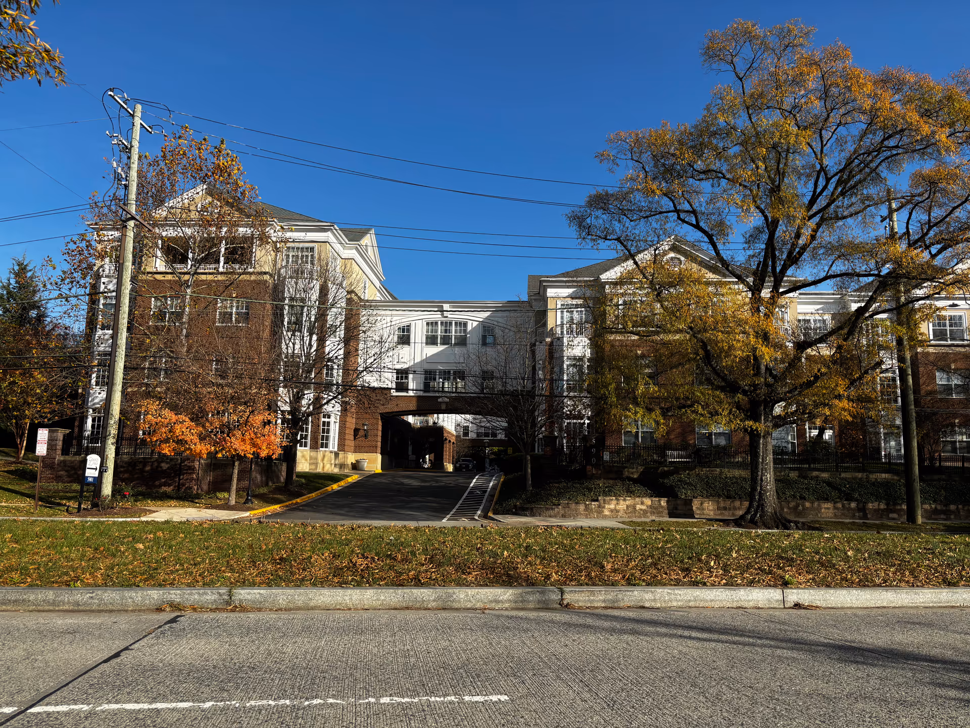Exterior view of a multi-story assisted living community building with brick and light-colored siding, surrounded by trees with autumn foliage under a clear blue sky. A driveway leads through an archway in the center of the building.