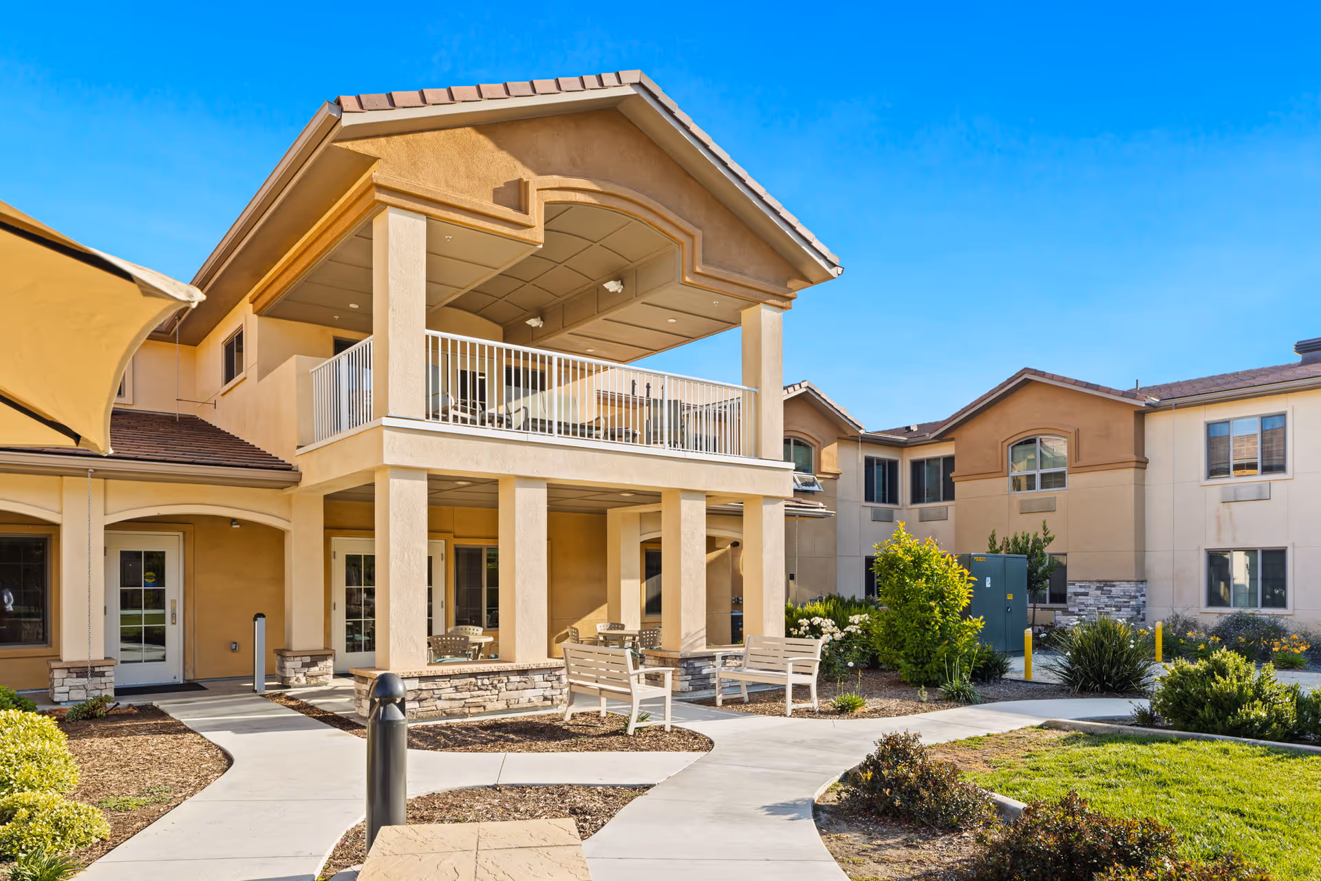 Exterior view of a two-story senior living facility building with beige stucco walls and a tiled roof under a clear blue sky. The building features a covered balcony with white railings on the second floor and a covered patio area with benches and chairs on the ground floor. There are paved walkways, landscaped bushes, and green grass surrounding the building.
