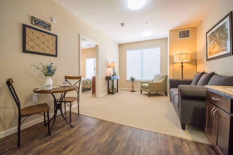 A cozy living area in a senior living facility featuring a small round wooden table with two chairs on a wood floor, a beige carpeted seating area with a green armchair, a dark gray sofa, two floor lamps, a small side table with a plant, and a framed picture on the wall. A doorway leads to a bedroom with a bed visible in the background.