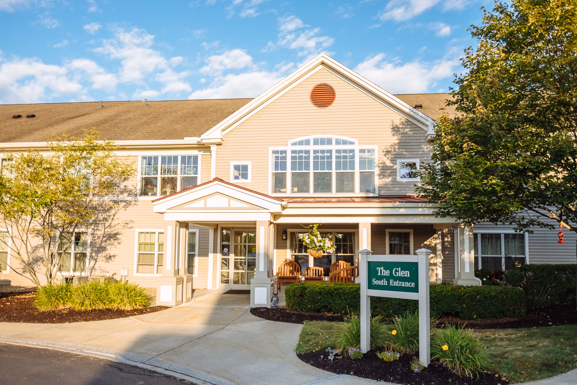 Exterior view of The Glen at Hiland Meadows showing the south entrance of a beige two-story building with white trim, a covered porch with wooden chairs, surrounded by trees and landscaping under a partly cloudy blue sky.
