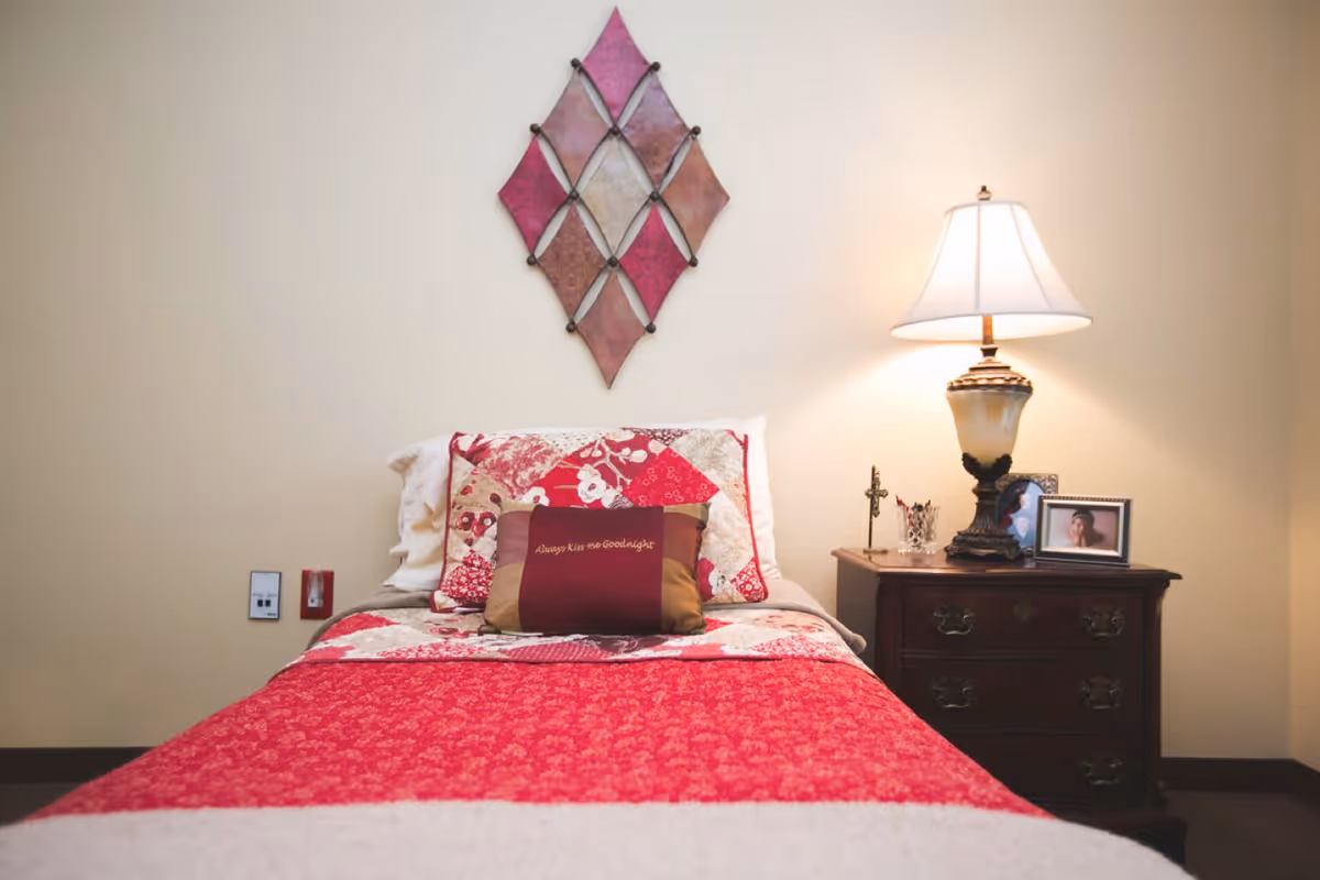 Single bed with a red quilt and decorative pillows, a nightstand with a lamp and framed photo, and wall art above in a bedroom.