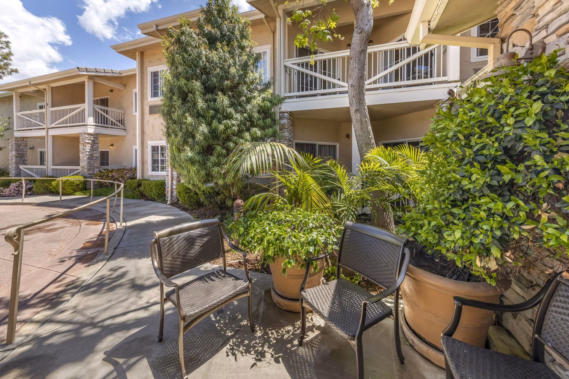 Outdoor seating area at Meadowbrook At Agoura Hills featuring metal chairs arranged around large potted plants with lush greenery, adjacent to a beige building with balconies and stone pillars under a partly cloudy sky.