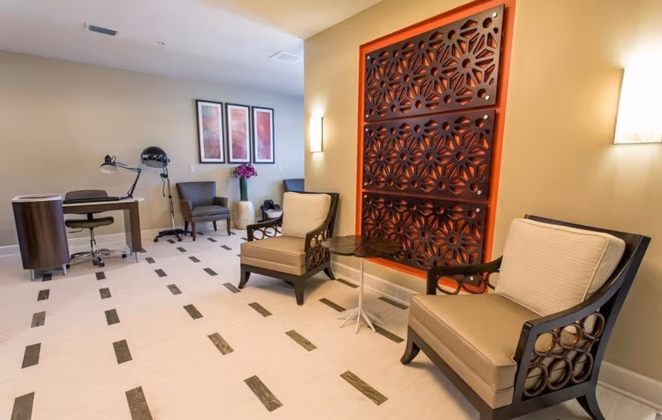 A well-lit interior room with two cushioned armchairs featuring wooden armrests and a small round side table between them. The wall behind the chairs has a large decorative wooden panel with a floral pattern. In the background, there is a desk with a chair and a desk lamp, two additional chairs, a tall vase with flowers, and three framed artworks on the wall. The floor has a light color with dark rectangular inlays.