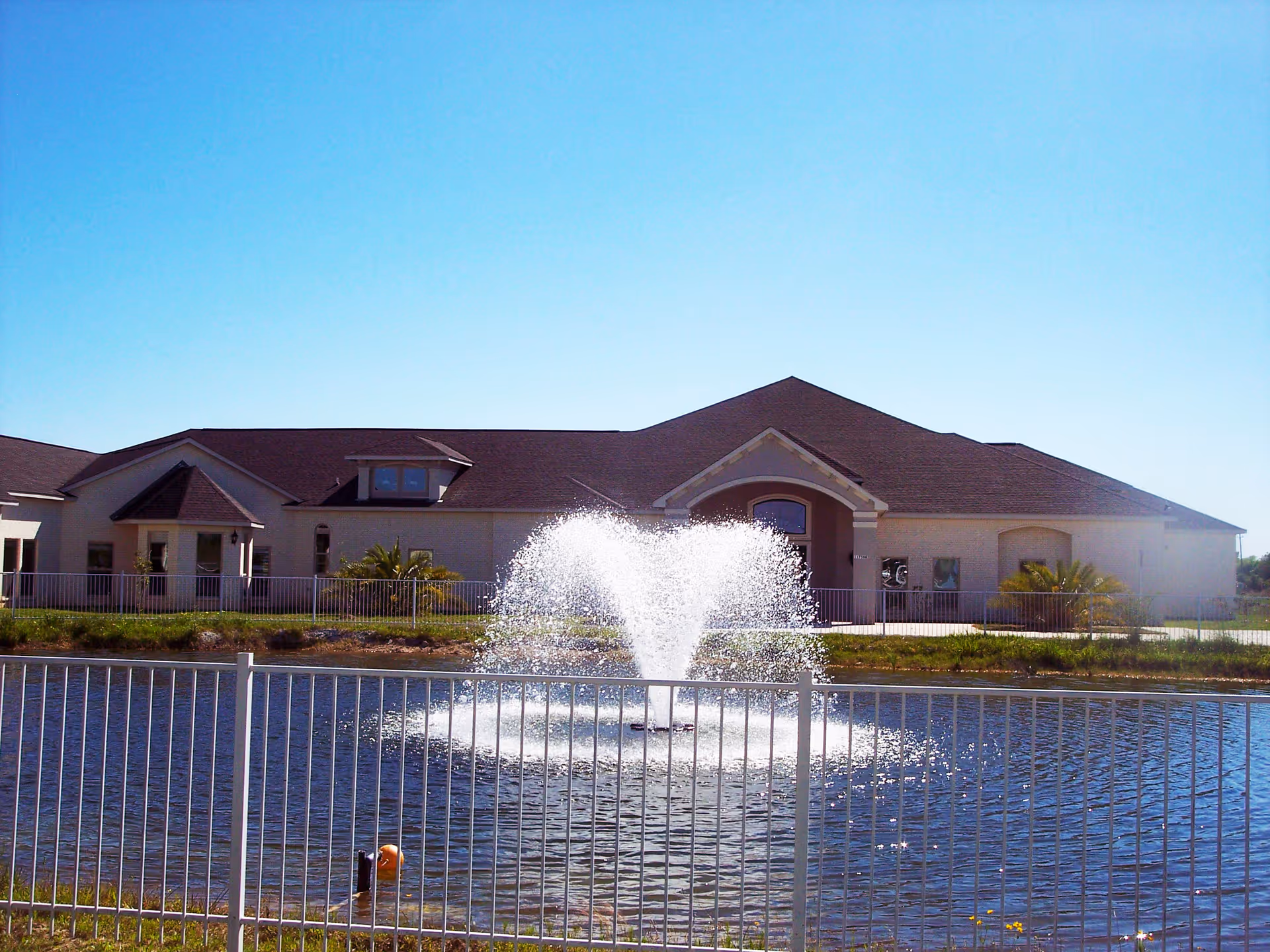 Single-story facility building viewed across a pond with a central water fountain and a metal fence in the foreground.