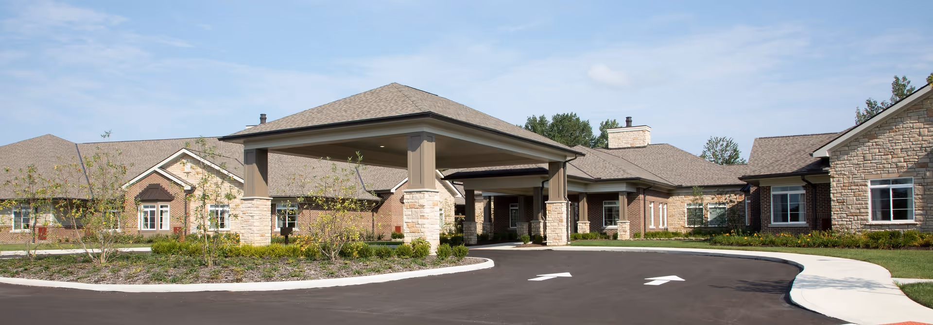 Exterior view of The Ganzhorn Suites senior living facility showing a large covered entrance with stone pillars, surrounded by landscaped greenery and a paved driveway with directional arrows.