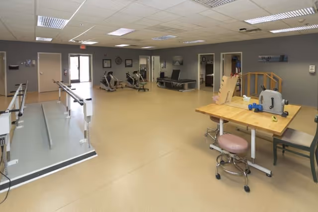 A spacious rehabilitation room with exercise equipment including parallel bars for walking practice, stationary bikes, and a therapy table. There is a wooden table with therapy tools and chairs on the right side. The room has beige flooring, gray walls, and a drop ceiling with fluorescent lighting.