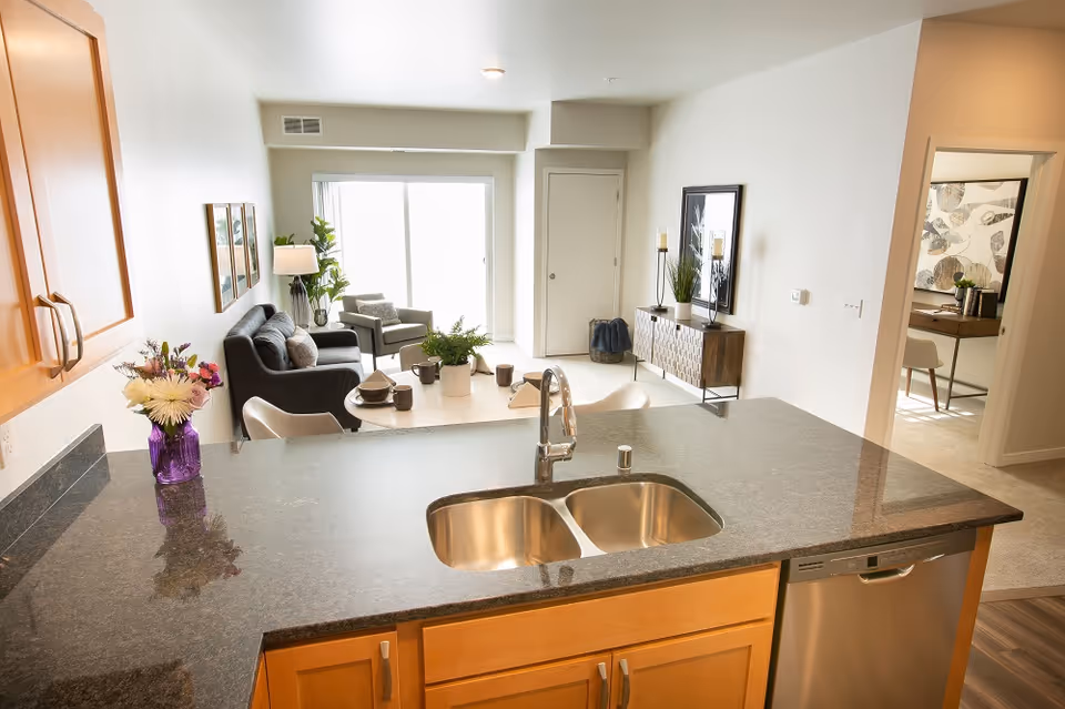 Kitchen island with double sink and granite countertop overlooking a bright living room with sofa, dining table, and sliding glass door.