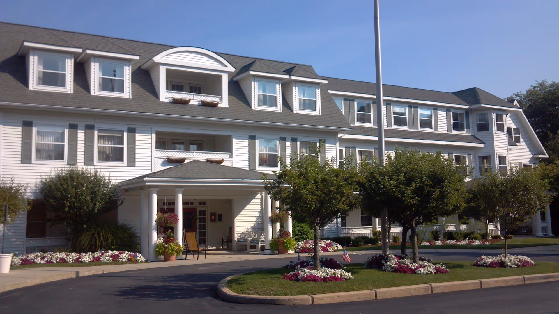 Exterior view of a three-story senior living facility with white siding and multiple windows. The entrance has a covered porch with columns, potted plants, and outdoor seating. The landscaped area in front features small trees and flower beds with red, white, and purple flowers. The sky is clear and blue.