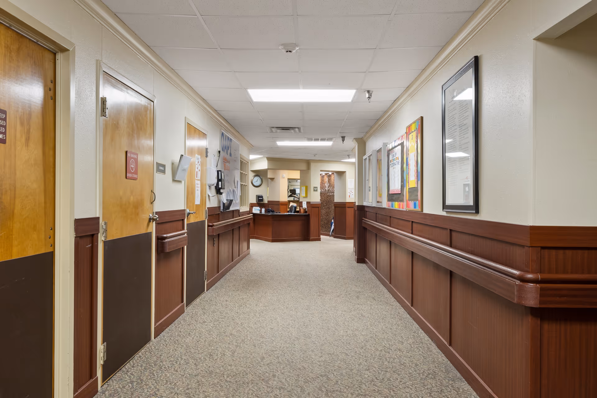 A well-lit hallway in a senior living facility with beige walls, wood paneling, and carpeted floor. Several wooden doors line the left side, some with signs, and bulletin boards and framed documents are displayed on the right wall. At the end of the hallway is a reception desk area with office equipment and a clock on the wall.