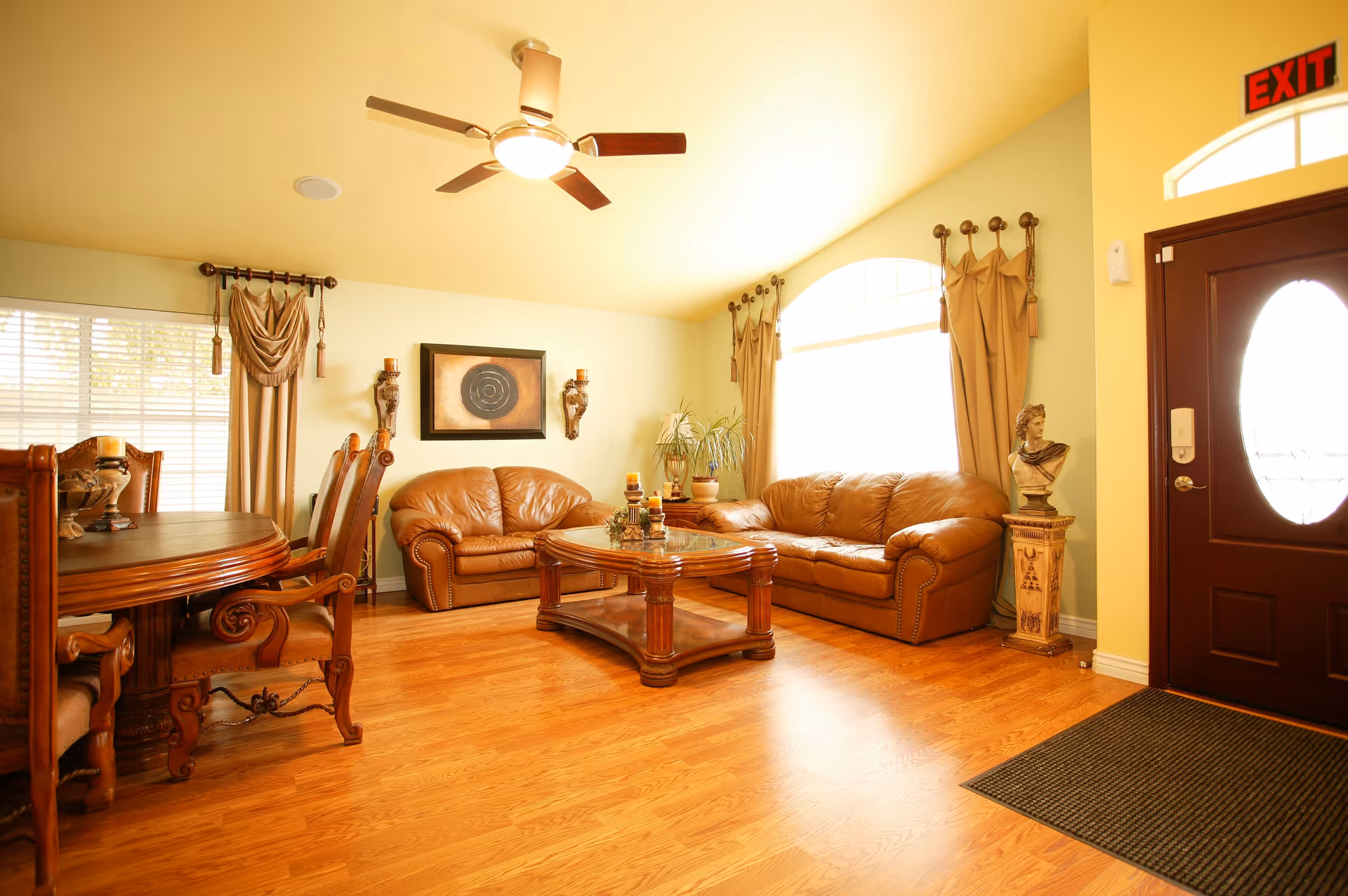 A warmly lit living room with wooden flooring, two brown leather sofas, a wooden coffee table with candles, a wooden dining table with chairs, a ceiling fan with light, large windows with drapes, a dark wooden front door with an oval glass panel, and a bust statue on a pedestal near the door.