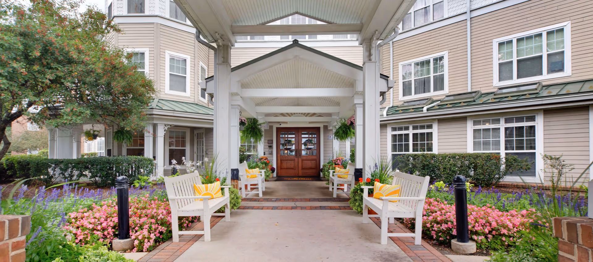 Covered main entrance of a senior living building with white benches, colorful flower beds, hanging plants, and double wooden doors.
