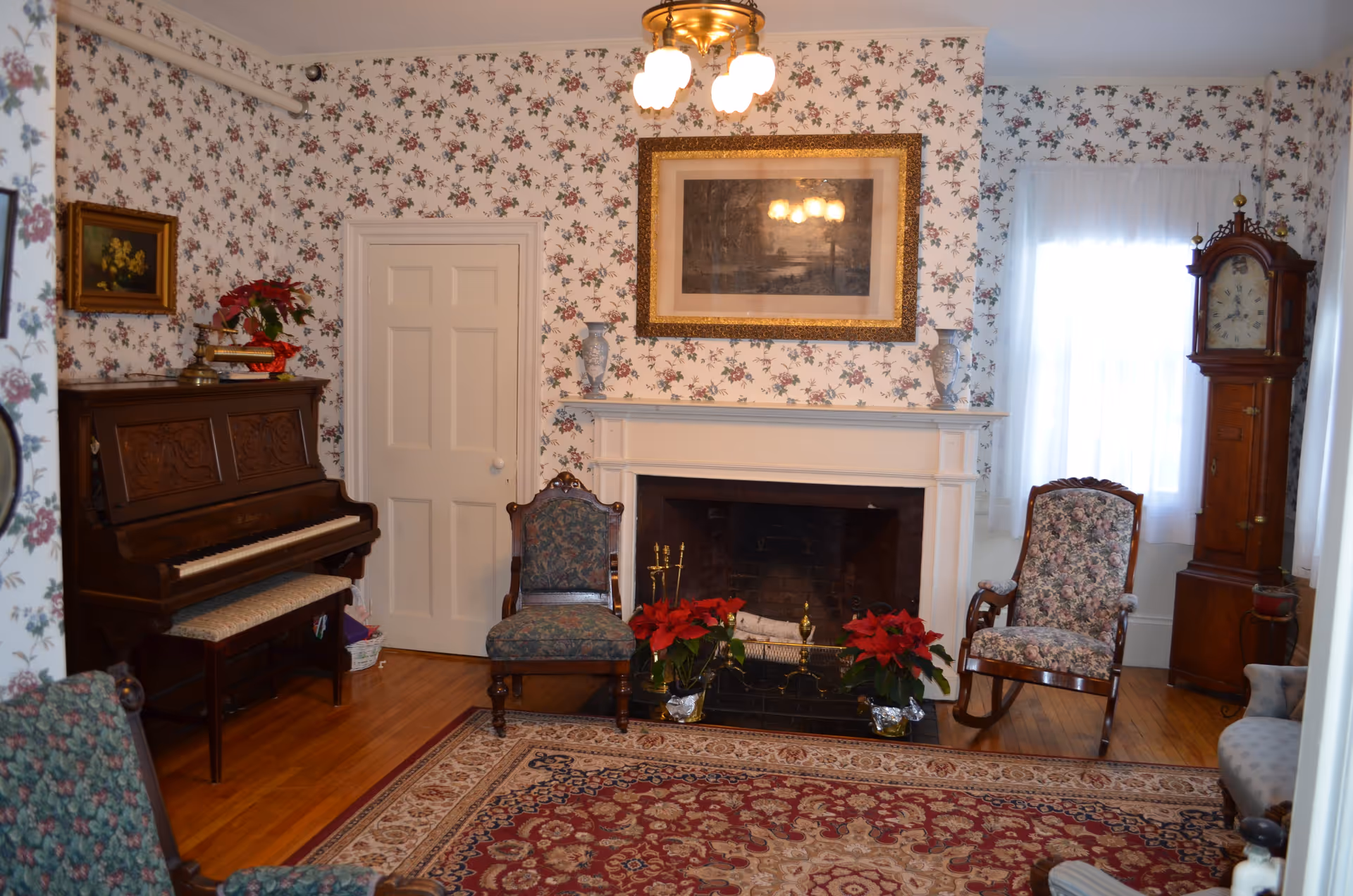 A cozy living room with floral wallpaper, a wooden piano with a cushioned bench, two upholstered chairs, a rocking chair, a grandfather clock, and a fireplace decorated with two red poinsettia plants and two blue vases. A framed picture hangs above the fireplace, and a chandelier with four lights is on the ceiling.