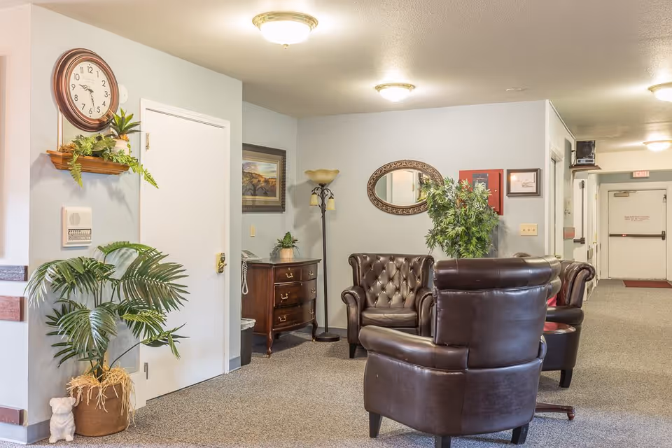 A cozy seating area in a senior living facility with three dark brown leather armchairs arranged around a small table. The walls are light-colored with a round mirror and framed artwork. There are several green plants, a wooden chest of drawers, a floor lamp, and a wall clock above a small shelf with decorative greenery. The carpeted floor extends down a hallway with doors and exit signs visible.