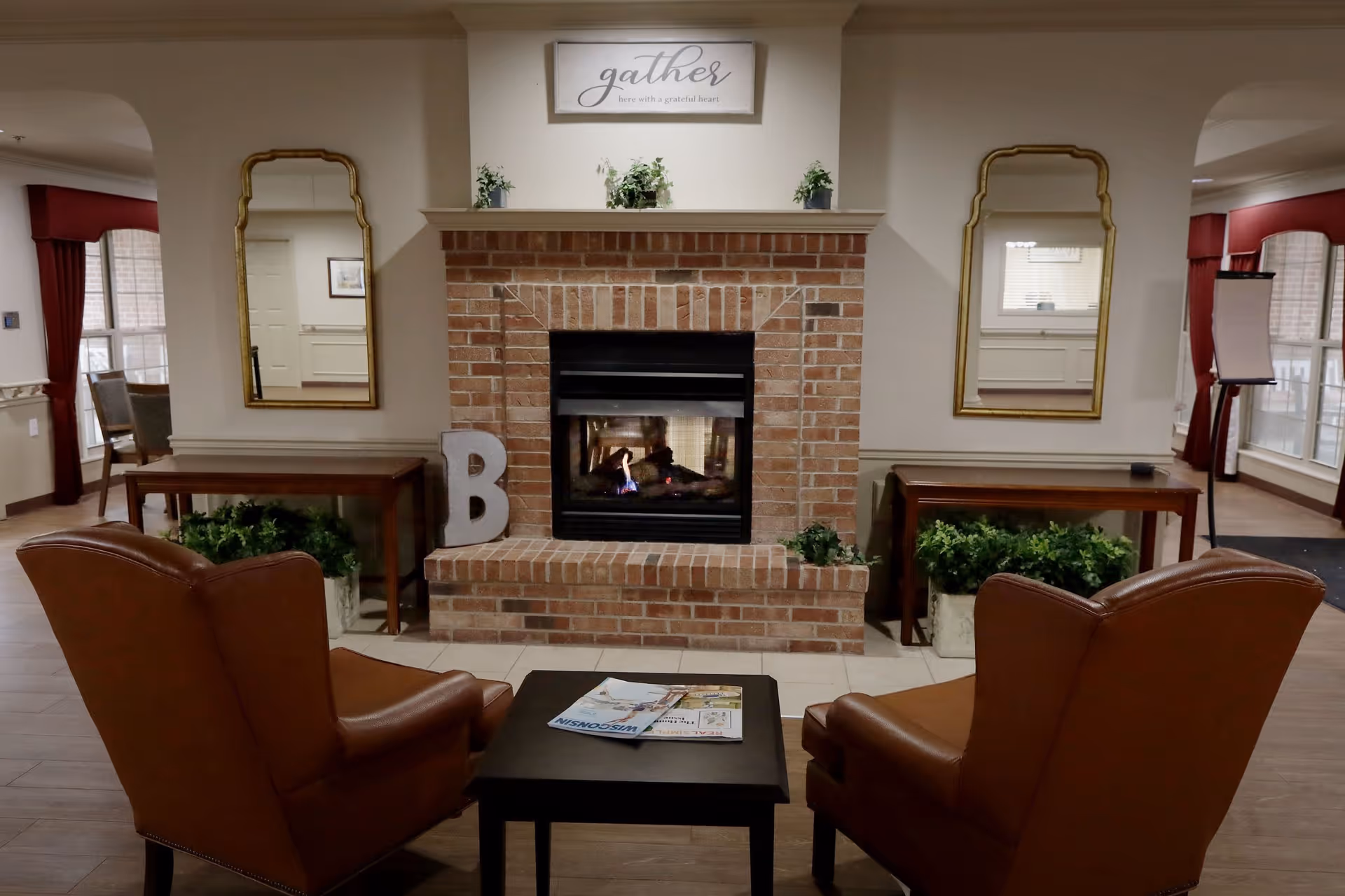 A cozy seating area in a senior living facility featuring two brown leather armchairs facing a brick fireplace with a small black coffee table in between. The fireplace mantel is decorated with small plants and a sign that reads 'gathers here with a grateful heart'. Two gold-framed mirrors hang on the wall on either side of the fireplace above wooden tables with green plants underneath. The room has large windows with red curtains and a wooden floor.